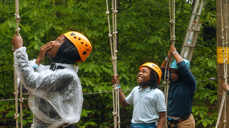Three young people wearing helmets are participating in a ropes course. The person on the left is wearing a clear plastic poncho and has their hand up to their face. The person in the middle is smiling and looking up. The person on the right is wearing a blue shirt and is also looking up. The ropes course is set in a wooded area.